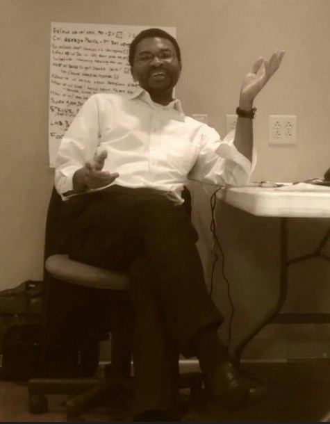 Dennis Rookwood in 2013, seated beside a folding table in a hospital consulting room, gesturing toward a flip chart covered in handwritten notes